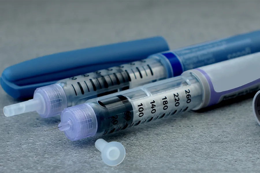 Two insulin pens with clear dosage windows and blue caps are placed on a gray surface, with a needle cover lying nearby.
