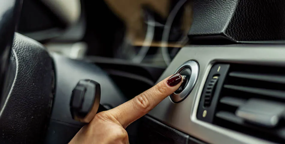 A person with dark red nail polish presses the engine start button on a car dashboard.