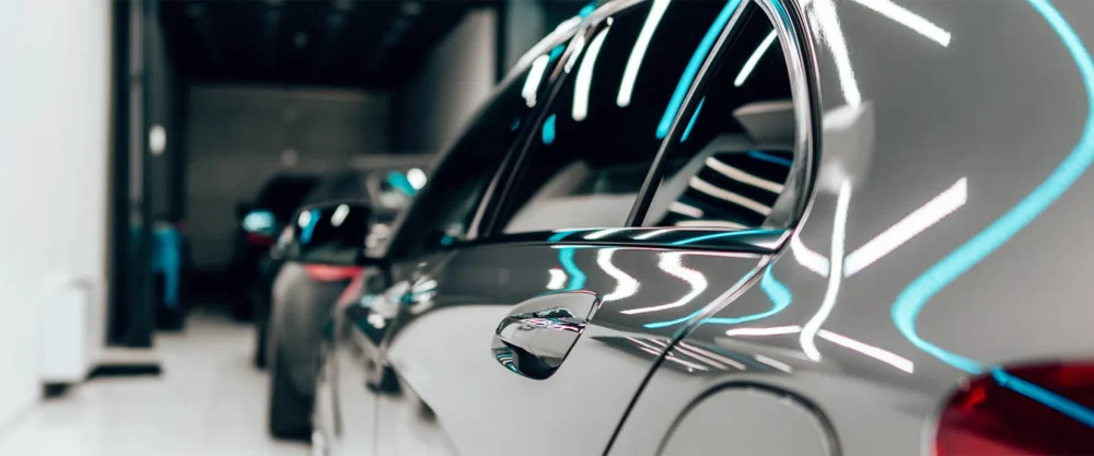 Close-up side view of a shiny gray car in a well-lit showroom, with reflections of lights on its surface and other vehicles in the background.