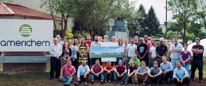 A group of people poses in front of an Americhem sign and building, some holding a large architectural rendering.