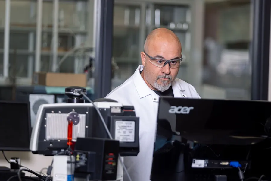 A researcher wearing safety glasses and a lab coat works at a computer in a laboratory setting with scientific equipment nearby.