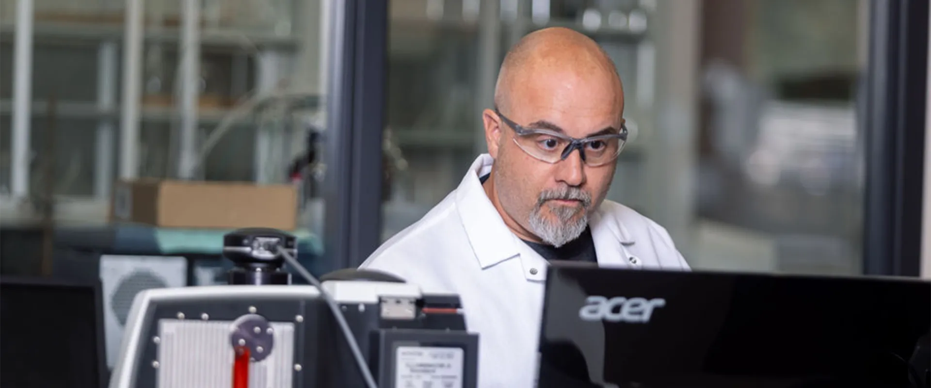 A man in a lab coat and safety glasses works at a computer in a laboratory setting.