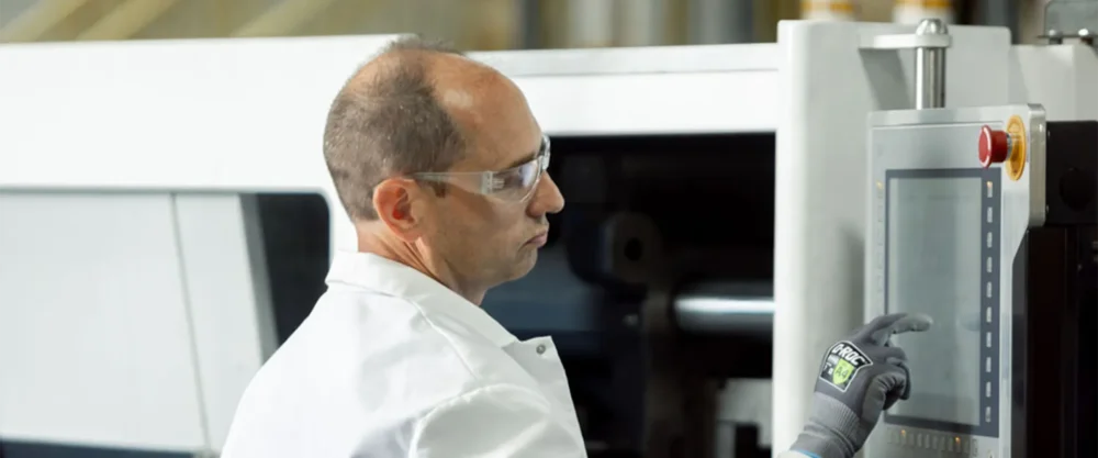 A person wearing safety glasses and gloves operates a touchscreen control panel on industrial machinery in a factory setting.