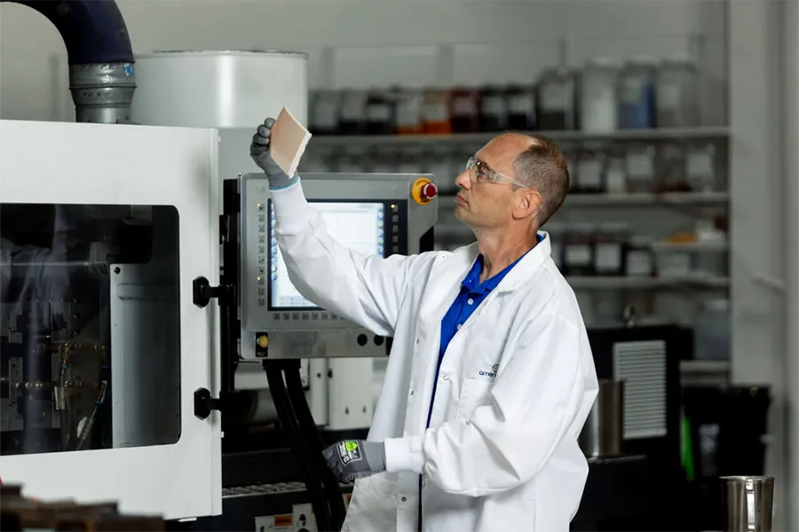 A man in a lab coat and gloves examines a sample in a laboratory or industrial setting with machinery and shelves in the background.