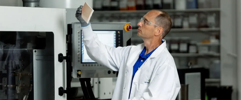 A scientist wearing a white lab coat and safety goggles examines a rectangular sample in a laboratory with equipment and shelves in the background.