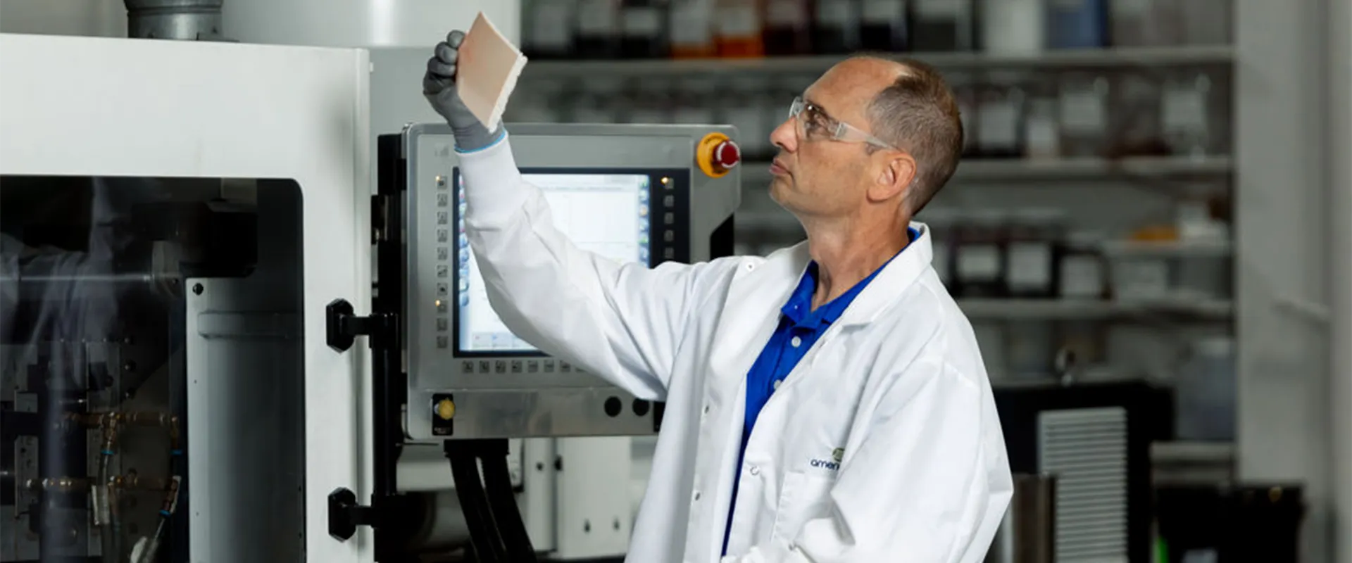 A scientist wearing a white lab coat and safety goggles examines a rectangular sample in a laboratory with equipment and shelves in the background.