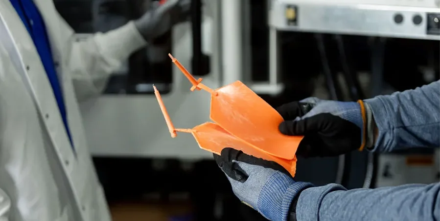 Person wearing gloves holds a broken orange plastic part, while another person in a lab coat stands nearby in an industrial or laboratory setting.