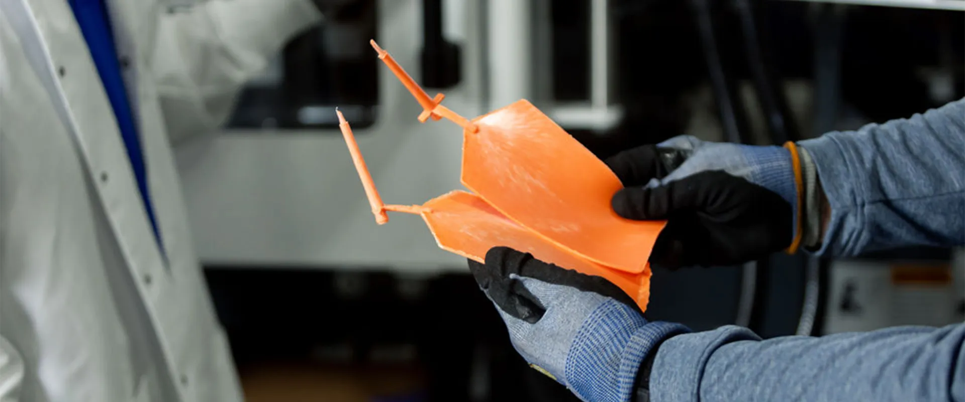 A person wearing gloves holds a large, orange, angular plastic part while another person in a lab coat stands nearby.
