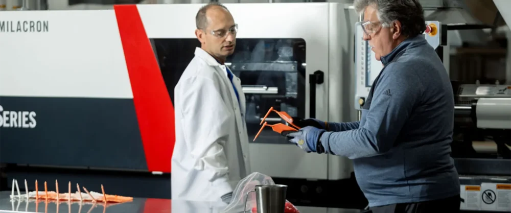 Two men wearing safety glasses examine a plastic part in a manufacturing facility with a large industrial machine in the background and several plastic items on a table.