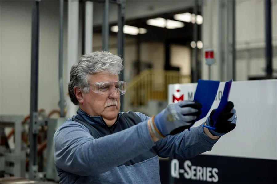 An older man wearing safety glasses and gloves examines two colored sheets in a factory or industrial setting.