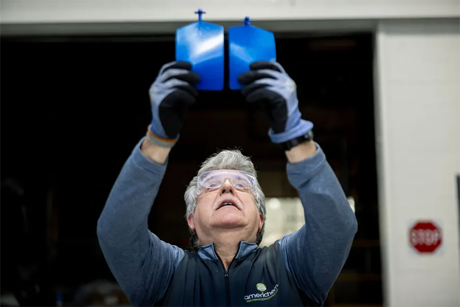 A person wearing gloves and safety glasses holds up two blue, flat objects for inspection inside an industrial setting.
