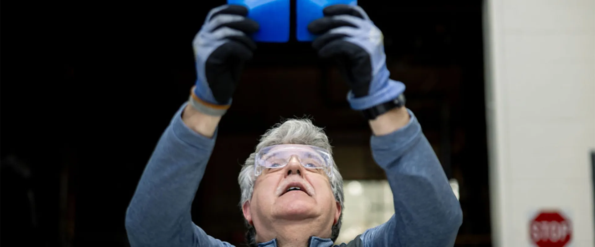 A person wearing safety glasses and gloves holds up a blue object, inspecting it closely with both hands raised above their head.