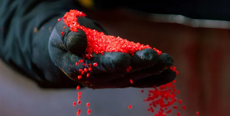 A gloved hand holds and pours small red plastic pellets.