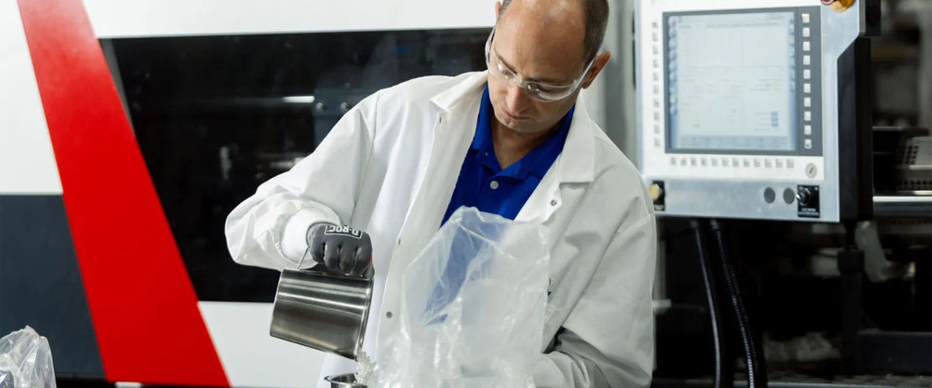 A person in a lab coat and safety glasses pours material from a metal container into a plastic-lined vessel in a laboratory with industrial equipment in the background.