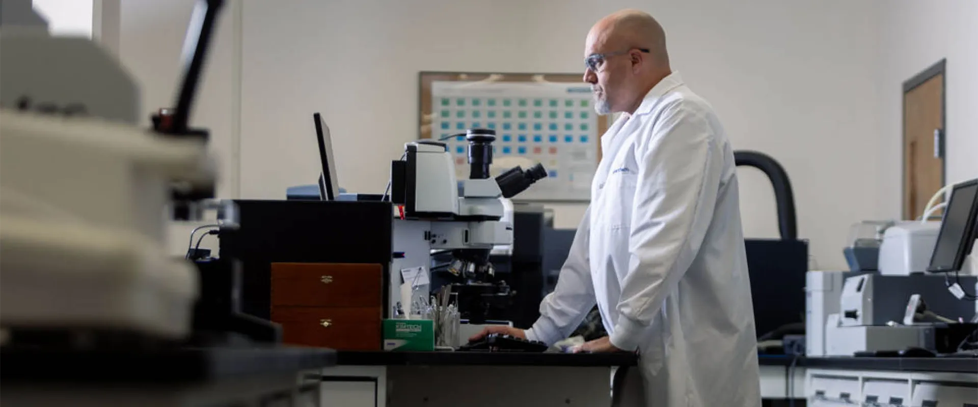 A scientist in a lab coat works at a computer and microscope in a laboratory setting.