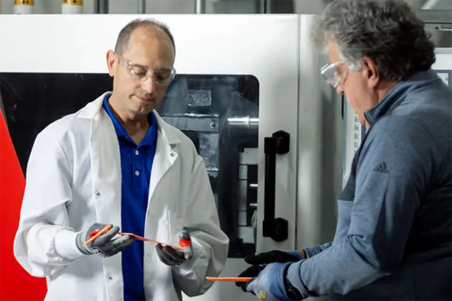 Two men wearing protective eyewear and gloves examine a red object in a laboratory or industrial setting with machinery in the background.
