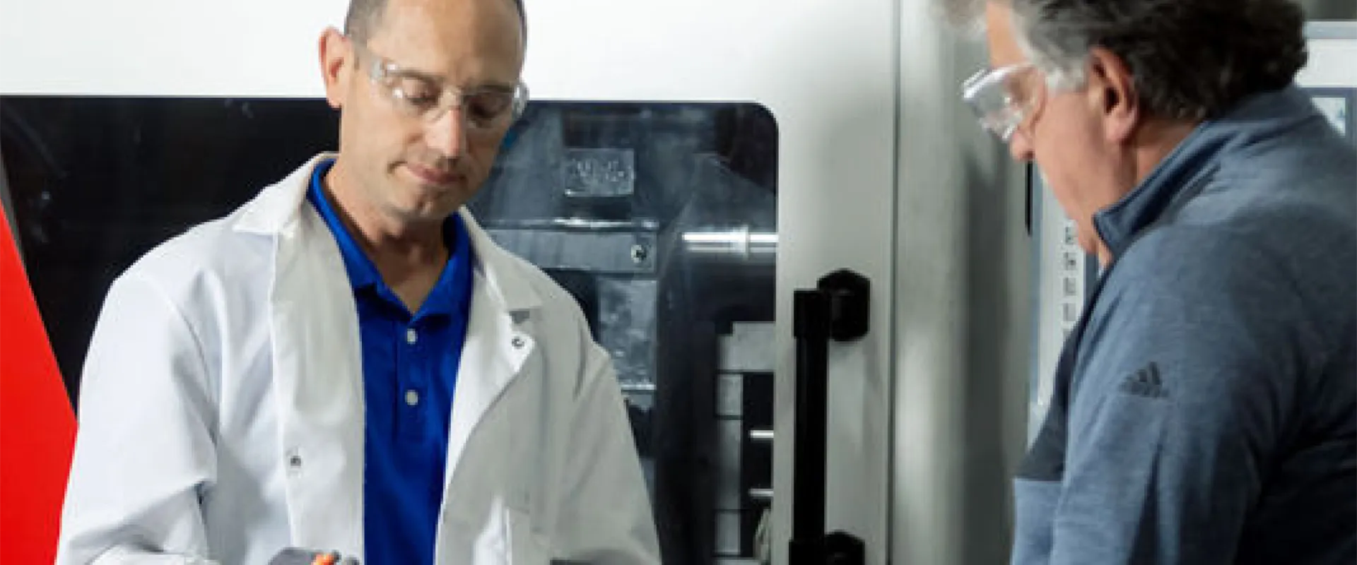 Two men wearing safety glasses examine a small object in a lab or industrial setting, with machinery visible in the background.