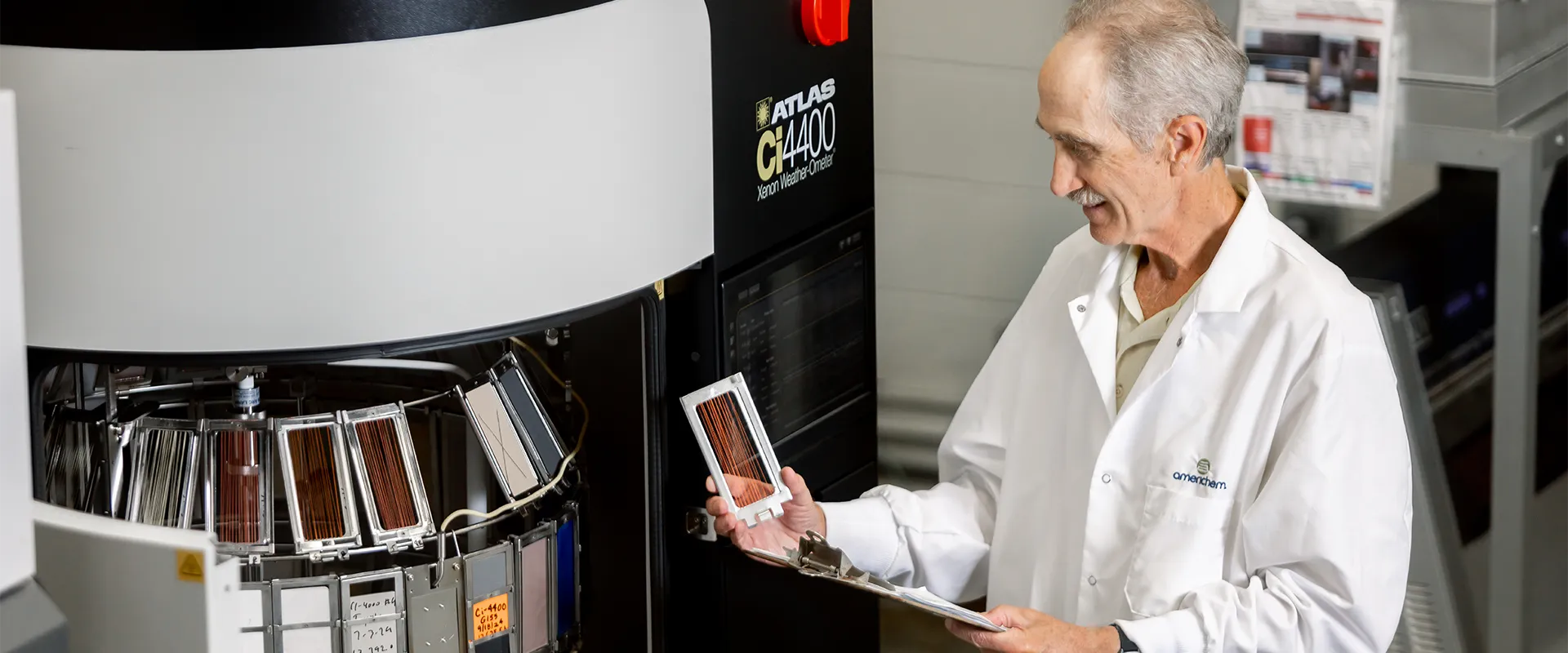 A scientist in a lab coat examines test samples in front of a large scientific instrument, holding a clipboard and a sample board.