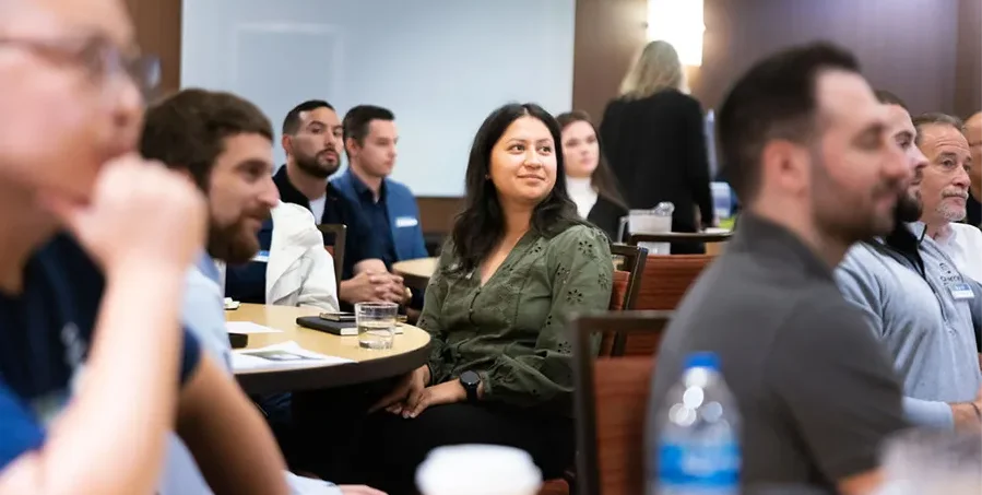 A group of people sit at round tables in a conference room, listening attentively. A woman in a green shirt is centered, looking forward and smiling slightly.