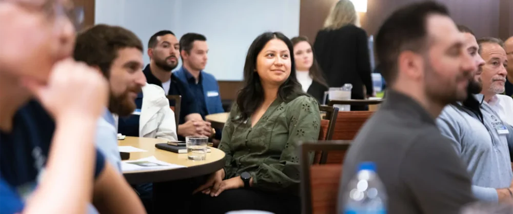 A group of people sit at tables in a conference room, listening attentively. One woman in a green blouse is looking forward and smiling.