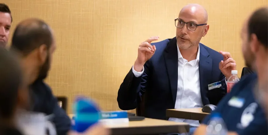 A man in a suit speaks and gestures with his hands during a meeting around a conference table with several other people.