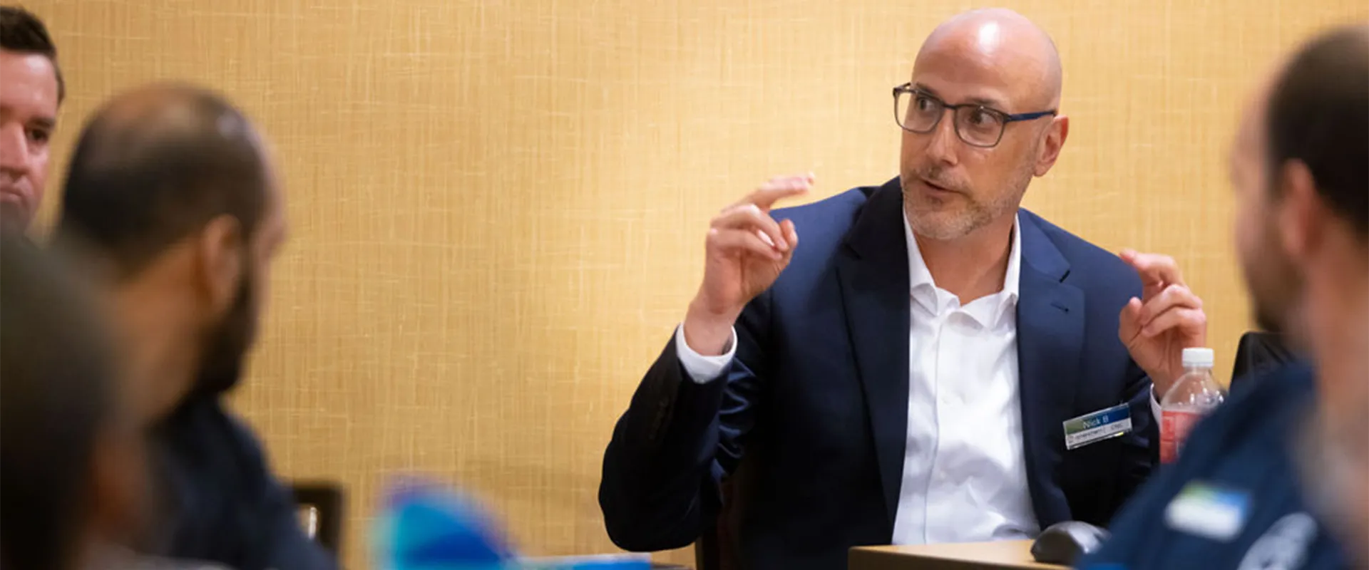 A man in a suit and glasses speaks while gesturing with his hands during a meeting at a conference table, with others listening.