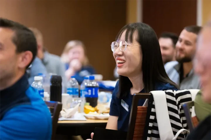 A group of people sits at tables with drinks and food, while a woman in glasses smiles and listens attentively in the foreground.