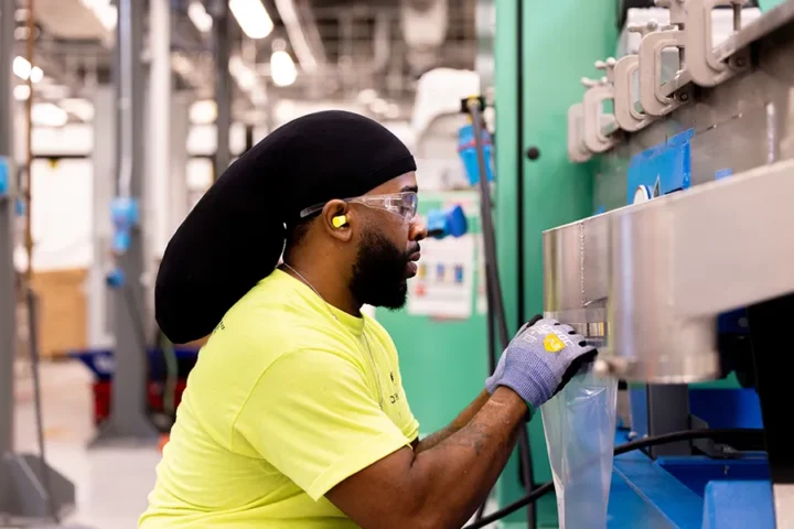 A worker in safety glasses, earplugs, and gloves operates machinery in an industrial facility.