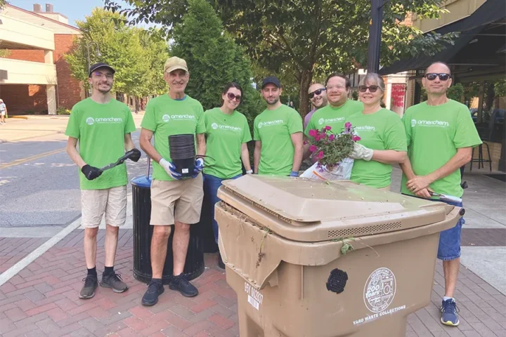 A group of seven people in matching green shirts stand outdoors on a sidewalk, holding gardening tools and pots near a trash bin and potted flowers.