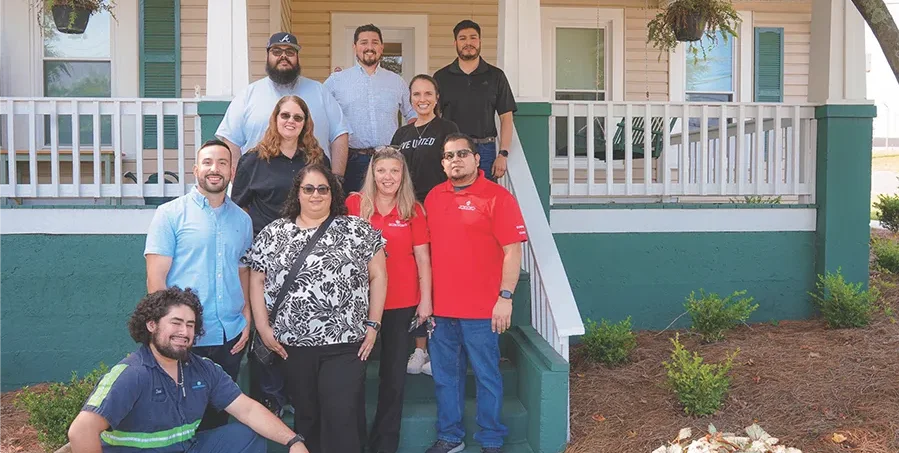 A group of ten people pose together on the steps of a porch in front of a house, smiling at the camera.