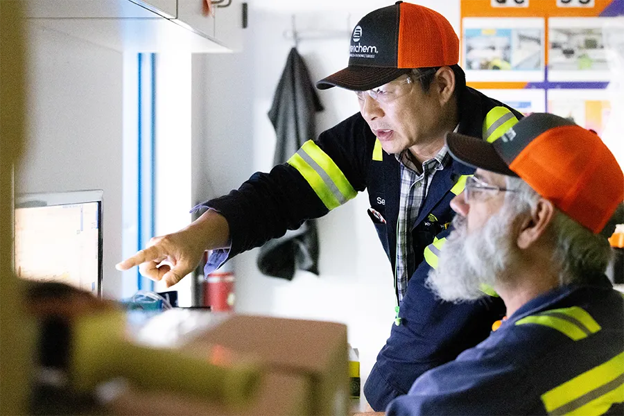 Two workers in safety gear look at a computer screen; one is seated while the other stands and points at the monitor, both wearing orange caps and reflective stripes.