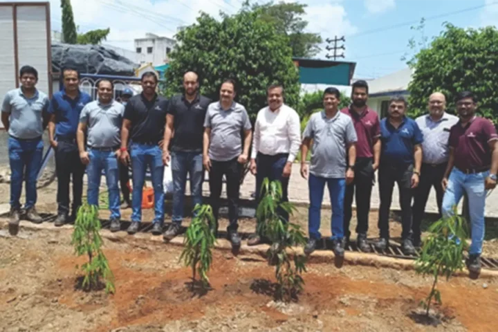 A group of people stands outdoors in a line behind several small, newly planted saplings on a sunny day.