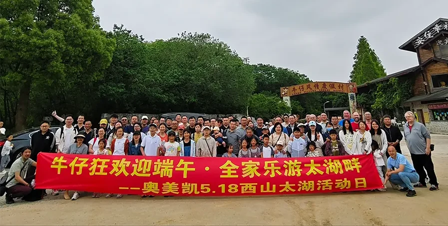 A large group of people of various ages pose outdoors, holding a red banner with Chinese text promoting a family outing event near a wooded area and rustic buildings.