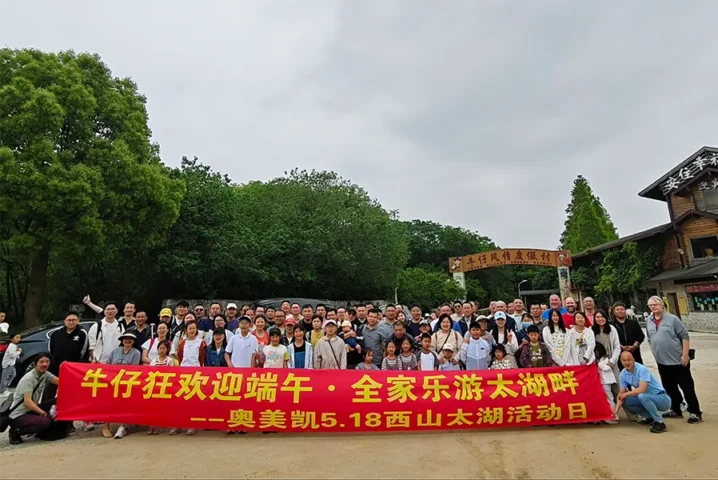 A large group of people poses outdoors holding a red banner with yellow Chinese text, with trees and a wooden building visible in the background.
