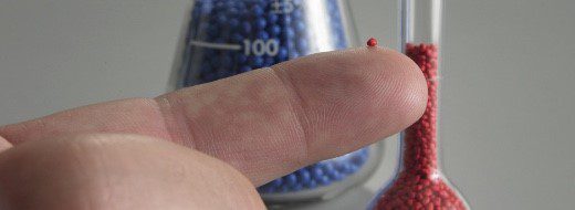 A small red bead rests on the tip of a finger, with blue and red beads in glass containers blurred in the background.
