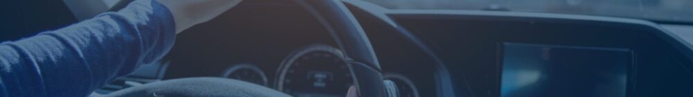 Close-up view of a person’s hand on a steering wheel, dashboard and speedometer visible in the background.