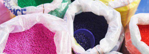 Open plastic bags containing colorful plastic pellets in shades of pink, green, dark blue, orange, and yellow are arranged closely together.