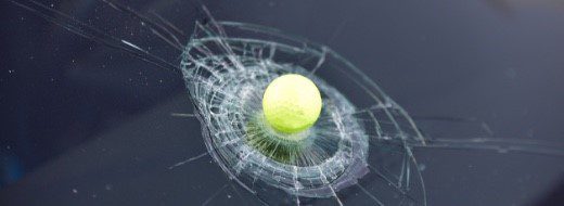 A tennis ball resting on a cracked car windshield, showing the impact point and shattered glass around it.