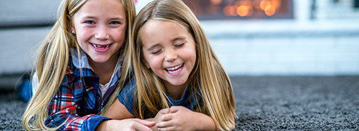 Two young girls with long blonde hair lie on a carpet, smiling and laughing together.