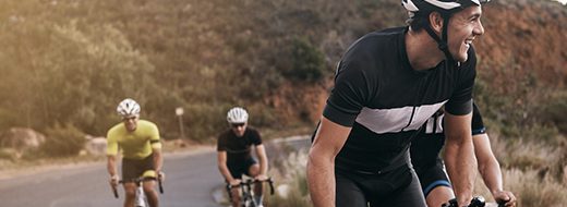 Four cyclists wearing helmets ride their bikes on a paved road through a scenic, natural landscape.