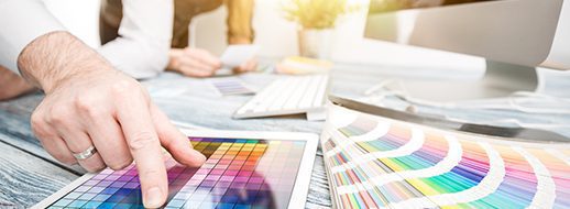 A person points at a color palette on a tablet while another works at a desk with a computer, color samples, and keyboards.