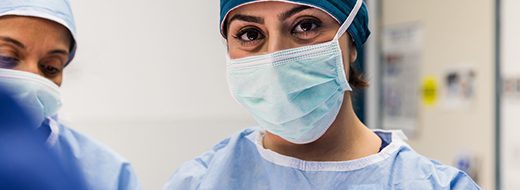 Two healthcare professionals wearing surgical masks, caps, and scrubs stand in a clinical setting, focusing on a task in front of them.