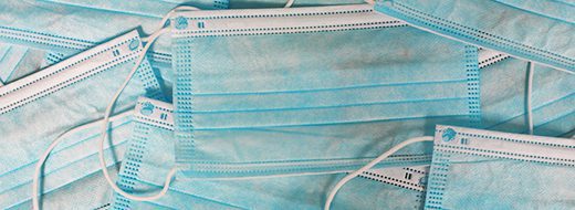 A pile of blue disposable surgical face masks overlapping each other, showing ear loops and pleated fabric.