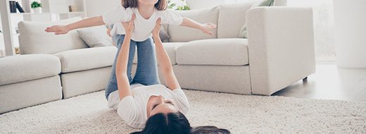 An adult lying on their back on a carpeted floor lifts a child in the air with their legs in a bright living room with a sofa in the background.