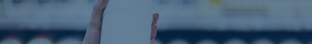 A hand holds a plain white cylindrical bottle against a blurred background of shelves.