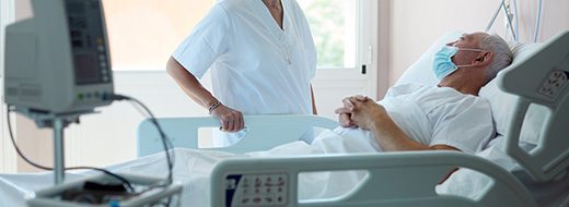 A nurse stands beside a masked elderly male patient lying in a hospital bed, with medical equipment visible in the foreground.