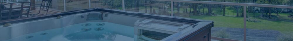 A hot tub on a deck with glass railings overlooks a green backyard and outdoor seating area.