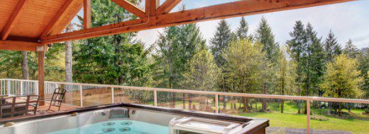 Covered outdoor patio with a hot tub, glass railing, and view of trees and a grassy yard.