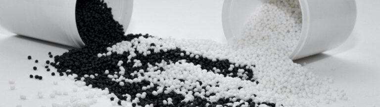 Two white containers tipped over, spilling black and white plastic pellets onto a white surface, with the colors mixing in the center.