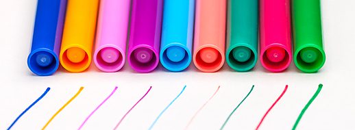 Eight colored markers arranged in a row with their caps facing forward, each drawing a matching colored line on a white surface.
